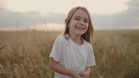 Ein-Glücklich-Lächelndes-Kleines-Mädchen,-Das-In-Die-Kamera-Blickt,-Läuft-Bei-Sonnenuntergang-In-Zeitlupe-In-Einem-Feld-Mit-Getreideähren